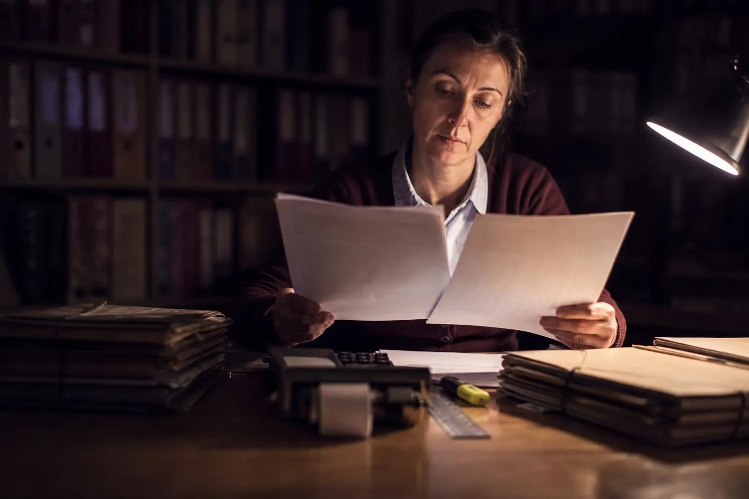 Une technicienne d’organisme de défense et de gestion compare deux liasses de déclarations papier sur une grande table de bureau éclairée par une lampe unique