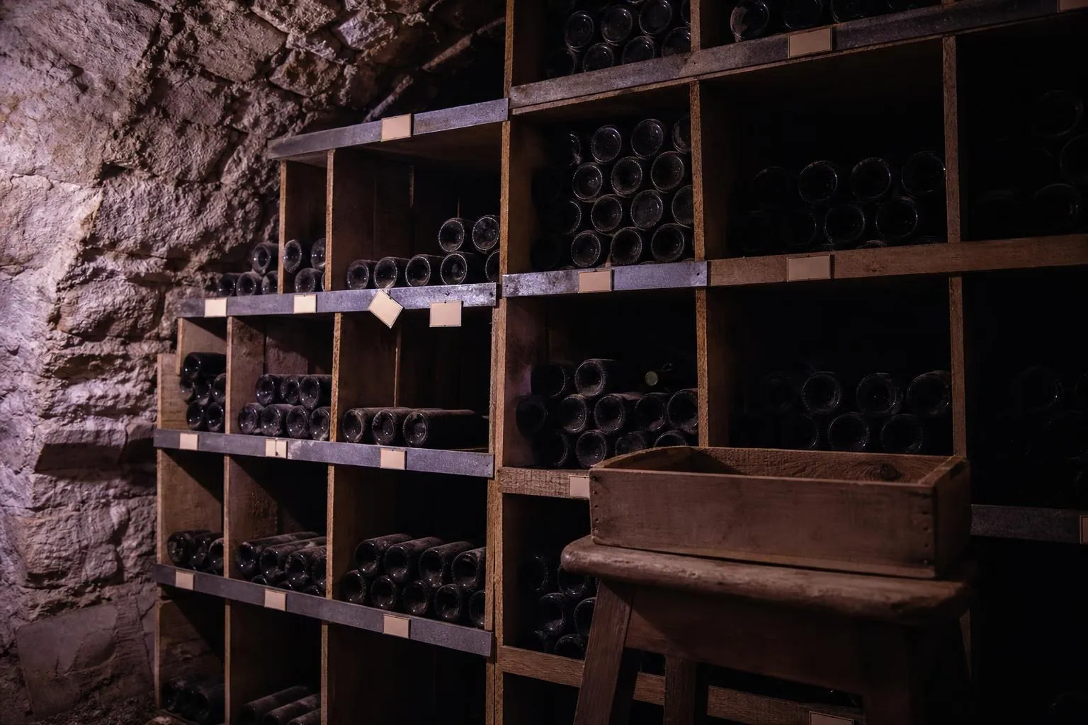 Mur de casiers en bois d’une cave, étiquettes de carton glissées dans des réglettes métalliques, certaines droites, d’autres de travers